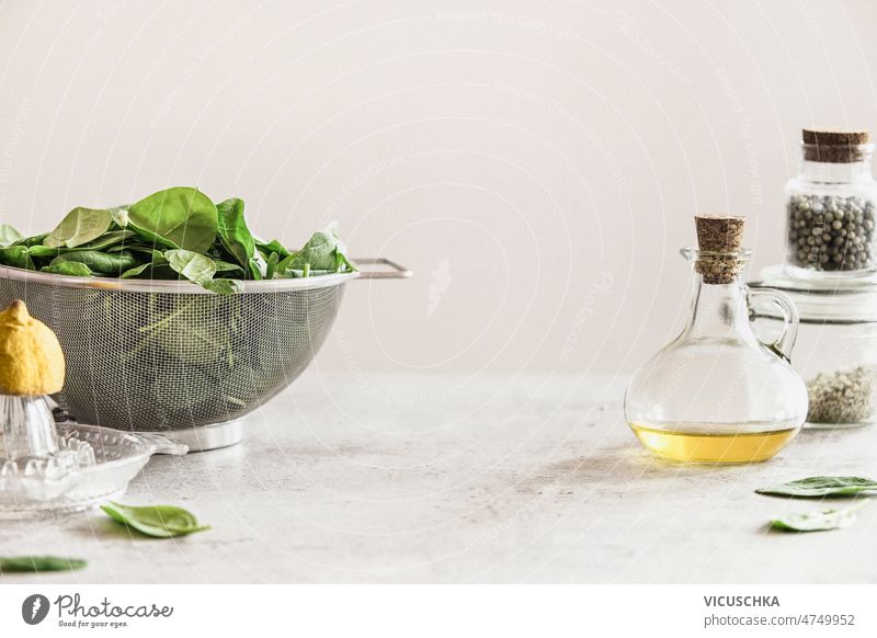 Food background with fresh spinach leaves in sieve, olive oil in glass bottle and citrus squeezer on white kitchen table at wall background. Healthy cooking at home. Front view.