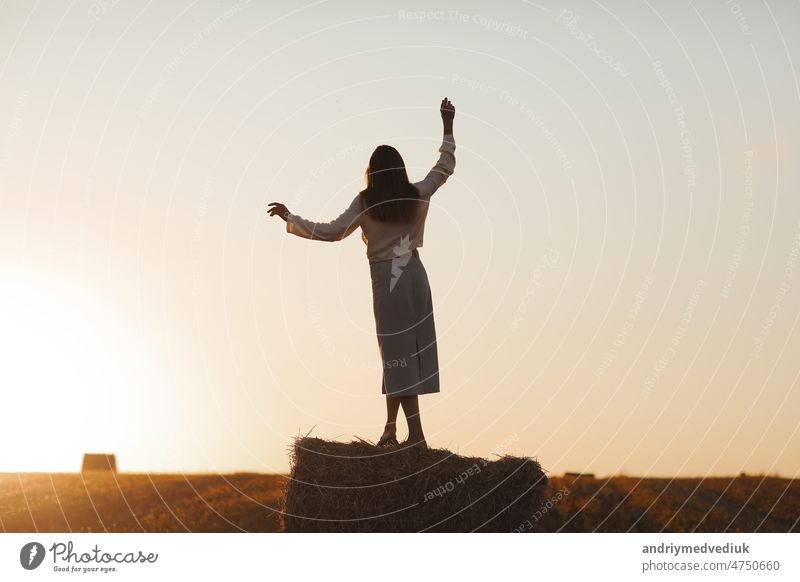 Young woman with long hair is standing and having fun on straw bale in field in summer on sunset. Female portrait in natural rural scene. Environmental eco tourism concept.