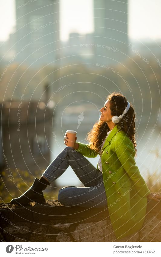 Pretty young woman listening music with smartphone and takeaway coffee by the river on a sunny autumn day pretty attractive caucasian outdoor beautiful