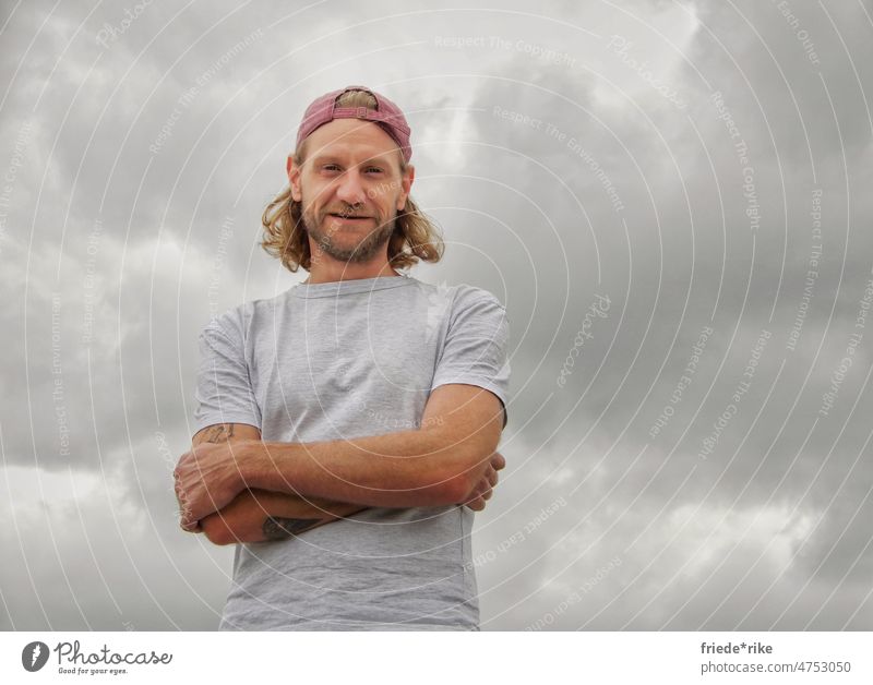 Man in front of cloudy sky portrait Facial hair Red Gray cappy Clouds Clouds in the sky Cloud cover crossed arms Curl Smiling Tattoo Blonde blue eyes skater