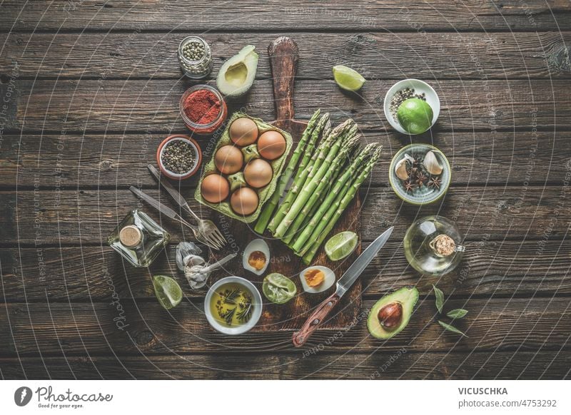 Various healthy cooking ingredients: green asparagus bunch, eggs, spices, avocado, lime,  olive oil various kitchen utensils forks knifes cutting board o