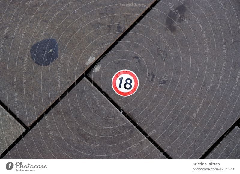 round sticker with red border and an 18 stuck on the sidewalk eighteen stickers forbidden interdiction off Ground of age Age of majority Sign symbol label Clue