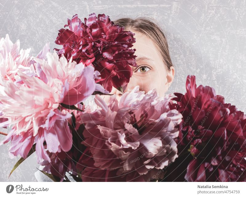 Large blurred bouquet of pink peonies covering face young woman on grey background. Focus are on one her eye. creative retro faceless focus on eye trend female