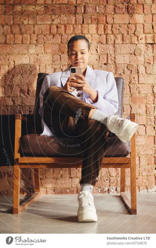 Portrait of confident multiracial LGBTQ mid adult woman sitting on retro chair and using smartphone in front of exposed brick wall portrait lgbtq business
