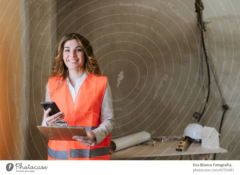 professional architect woman in construction site using mobile phone and blueprints. Home renovation workspace protective helmet protective jacket real estate