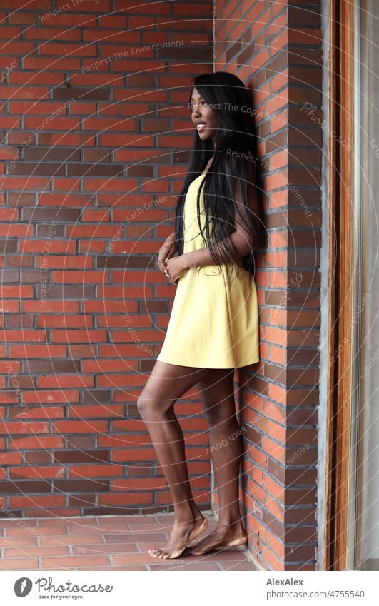 Young beautiful long haired woman in yellow summer dress stands barefoot on balcony in front of brick wall and smiles Woman Young woman pretty Beauty & Beauty