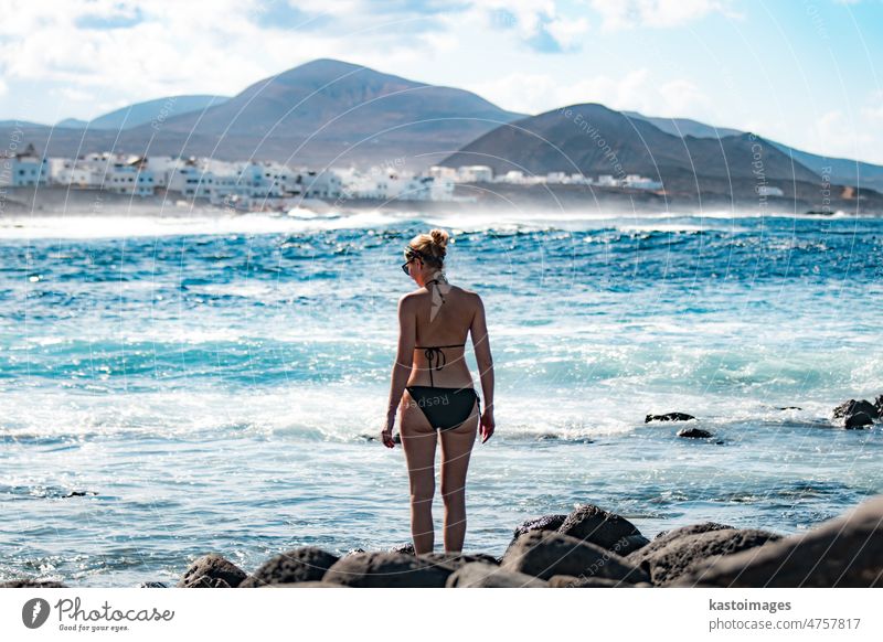 Female tourist at wild rocky beach and coastline of surf spot La Santa Lanzarote, Canary Islands, Spain. La Santa village and volcano mountain in background