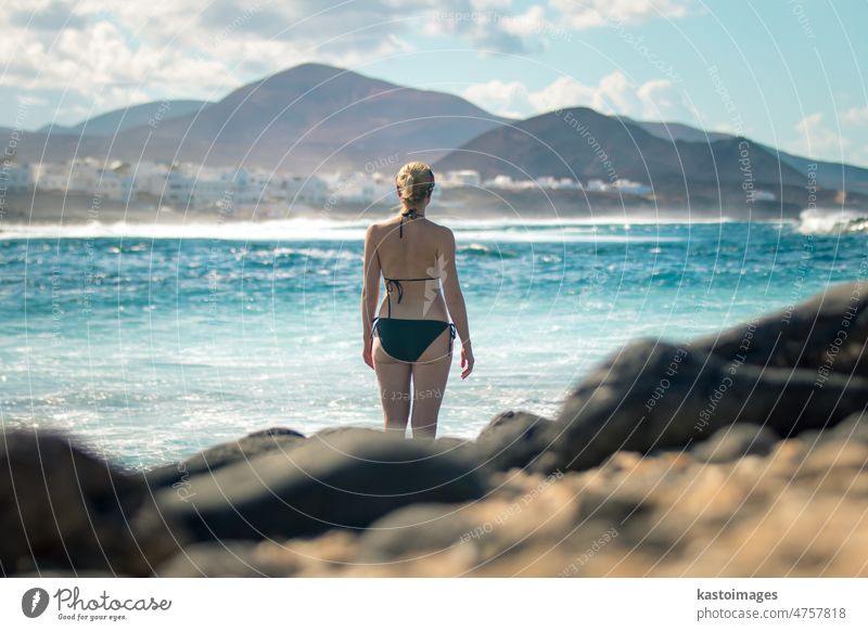 Female tourist at wild rocky beach and coastline of surf spot La Santa Lanzarote, Canary Islands, Spain. La Santa village and volcano mountain in background