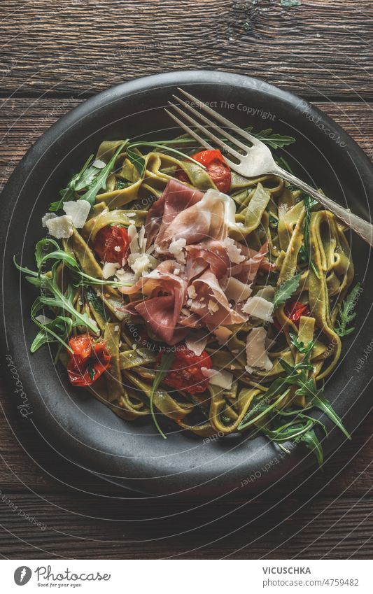 Close up of pasta dish with noodles, arugula, tomatoes, ham and cheese in black bowl close up fork rustic wooden kitchen table traditional italian food top view