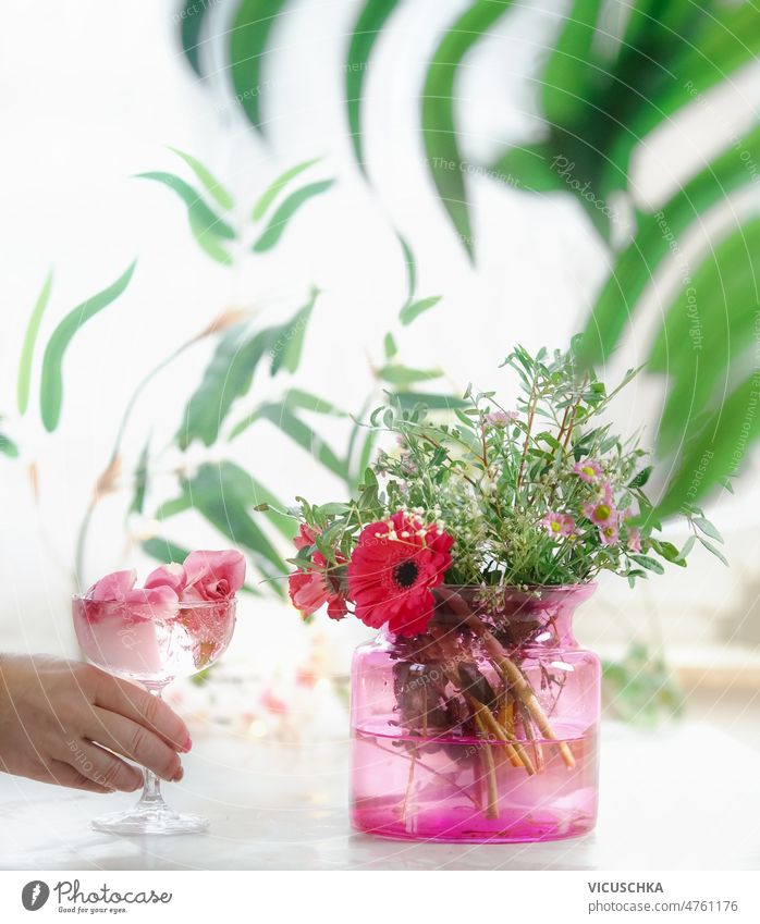 Women hand holding champagne glass with rose petals and flower ice cube on table with flowers bunch in pink glass vase women white blurred branch background