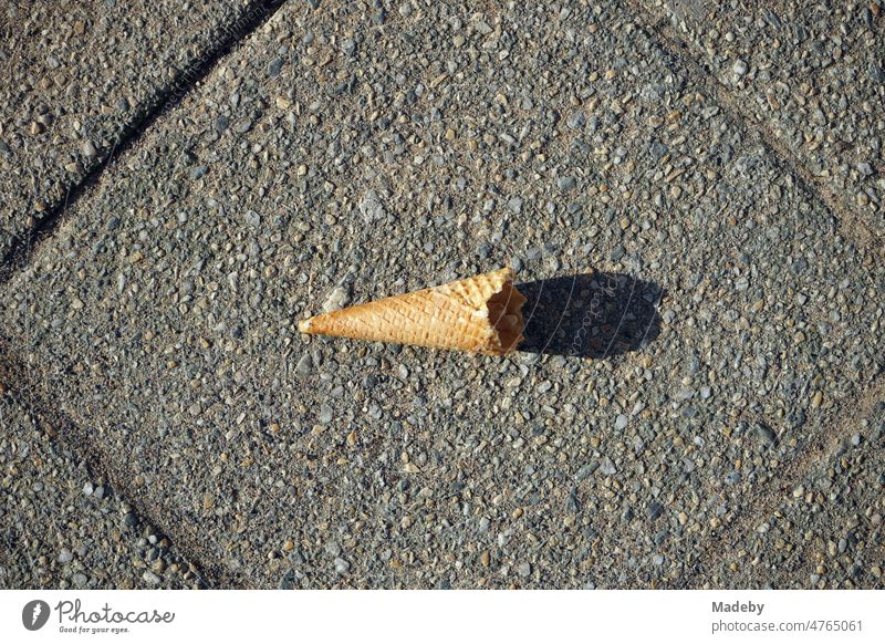 Empty nibbled wafer of an ice cream cone with light and shadow in sunshine on gray pavement in spring in the pedestrian zone in Detmold in East Westphalia-Lippe, Germany