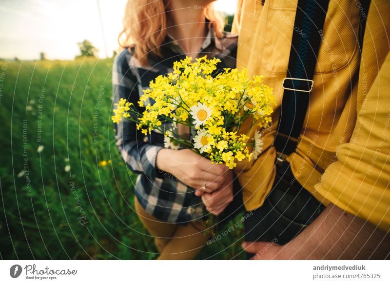 cropped photo of bouquet of wildflowers in woman's hand. Loving hipster couple walking in the field, kissing and holding hands, hugging, lying in the grass in the summer at sunset. valentines day