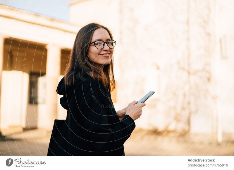 Smiling woman in eyeglasses and black hoodie using smartphone outdoors positive connection message millennial browsing city online female smile urban cellphone