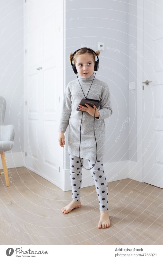 Curious girl with tablet listening to music in headphones using playlist digital addict free time depend device gaze curious gadget kid barefoot doorway pastime