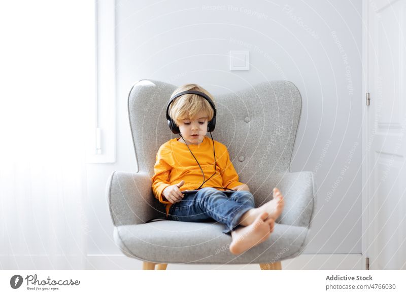 Barefoot boy in headphones watching cartoons on tablet listen using addict digital internet spare time home armchair interest kid browsing device movie online