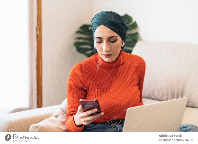 Muslim female using smartphone on sofa woman portrait laptop earphones earbuds tws wireless home rest listen music social media weekend muslim ethnic islam
