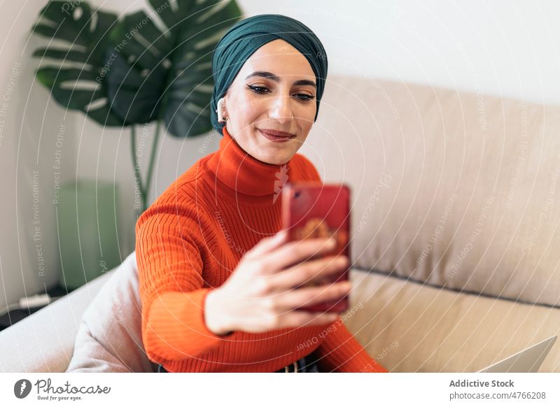 Muslim woman taking selfie at home portrait living room smartphone smile sofa music listen weekend female muslim ethnic islam headscarf gadget device turtleneck