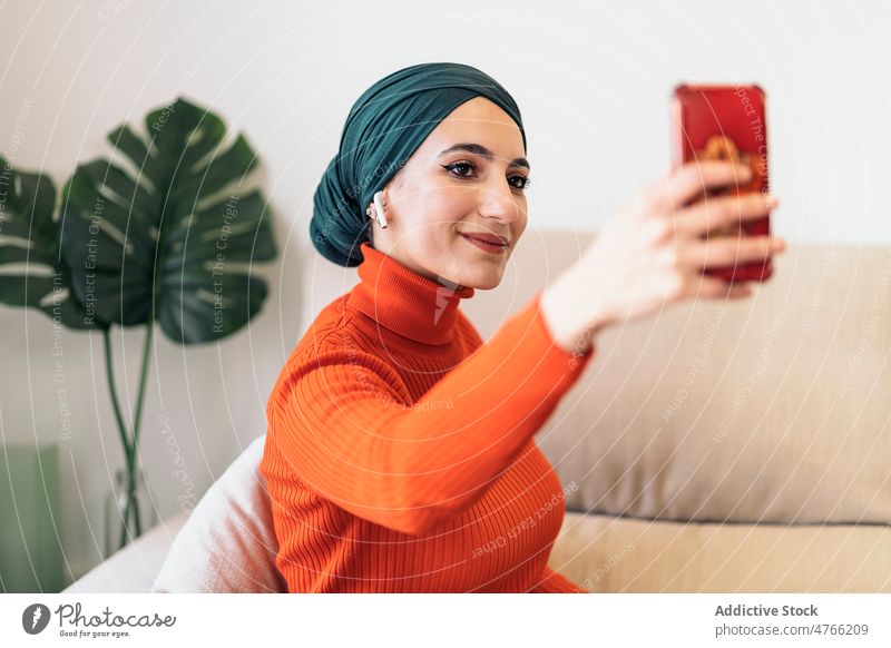 Muslim woman taking selfie at home portrait living room smartphone smile sofa music listen weekend female muslim ethnic islam headscarf gadget device turtleneck