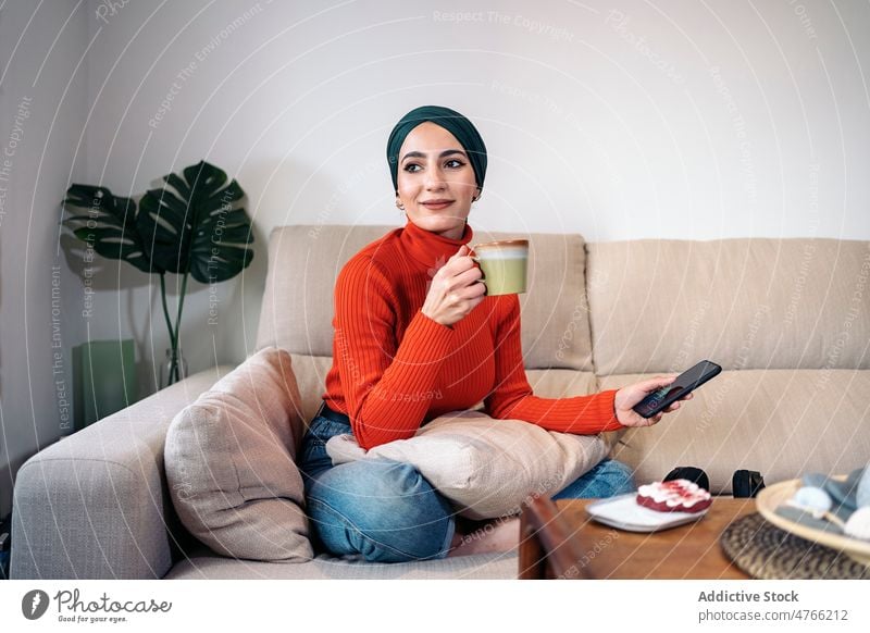 Muslim woman enjoying hot drink on sofa portrait rest weekend home smartphone smile living room glad female muslim ethnic islam legs crossed optimist tea coffee
