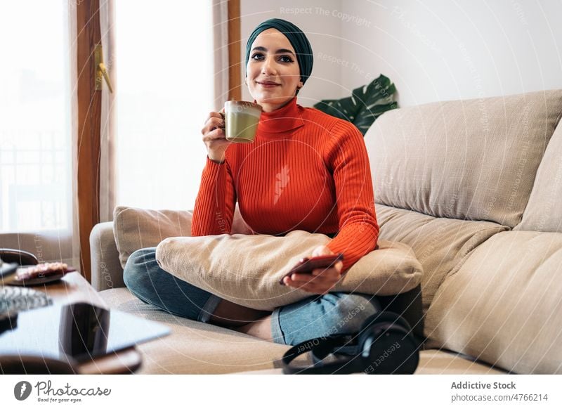 Muslim woman enjoying hot drink on sofa portrait rest weekend home smartphone smile living room glad female muslim ethnic islam legs crossed optimist tea coffee