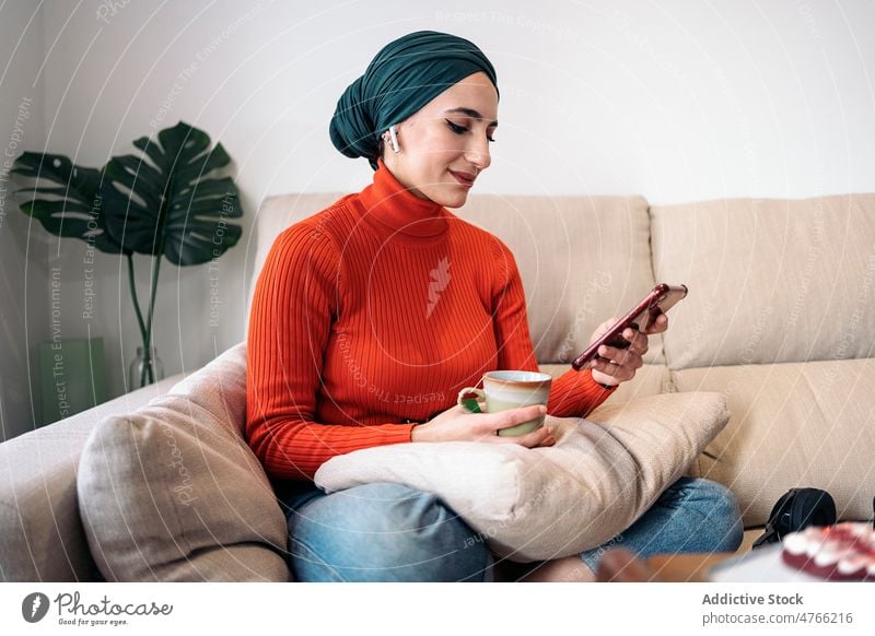 Muslim woman enjoying hot drink on sofa portrait rest weekend home browsing smartphone earphones earbuds smile wireless living room glad female muslim ethnic