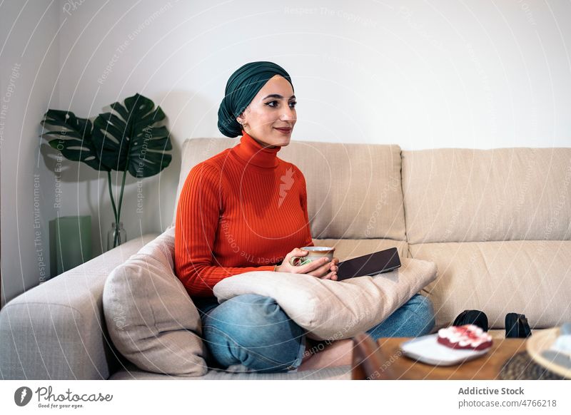 Muslim woman sipping hot drink portrait tea rest sofa weekend home living room daytime female muslim ethnic islam couch comfort headscarf cozy turtleneck