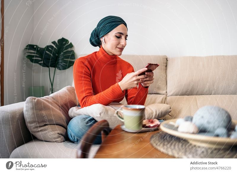 Muslim woman enjoying hot drink on sofa portrait rest weekend home browsing smartphone earphones earbuds smile wireless living room glad female muslim ethnic