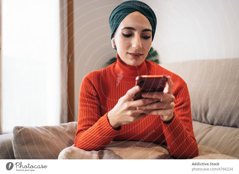Muslim female using smartphone on sofa woman portrait home rest earphones earbuds tws wireless listen music social media weekend muslim ethnic islam playlist