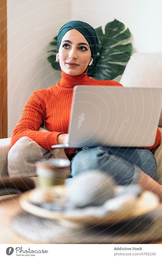 Muslim woman with laptop resting on couch portrait using sofa home earphones earbuds living room smile listen music female muslim ethnic islam online weekend