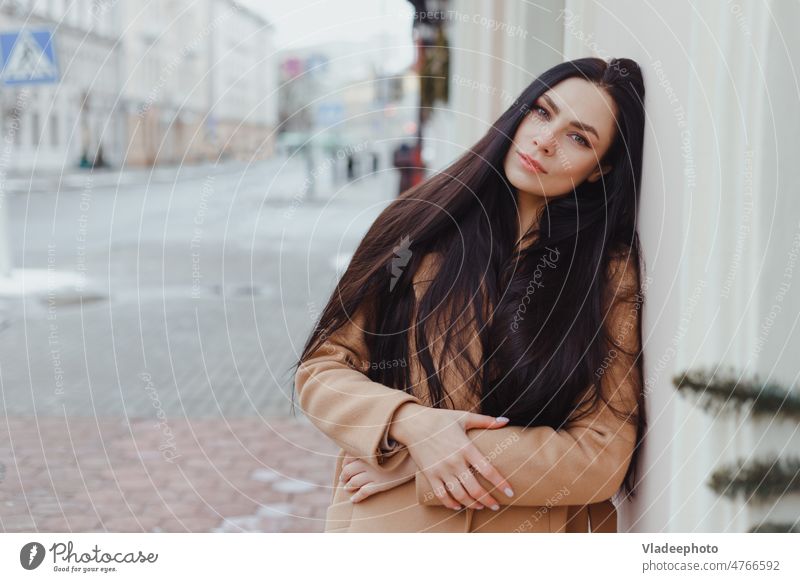Young brunette woman in beige winter coat stands alone on the street fashion clothes look elegant city long hair young wall beautiful lifestyle model white