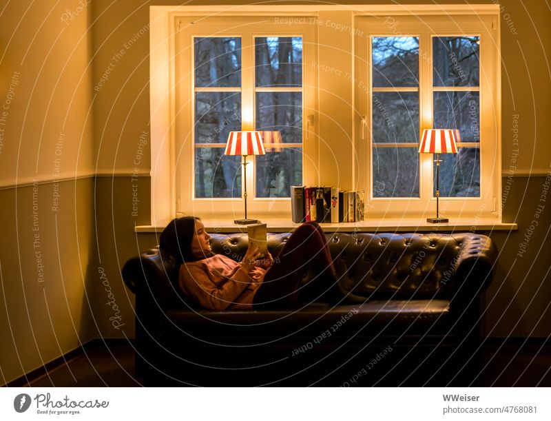 A young woman lies comfortably on a sofa by the window and reads, outside is blue hour Reading books Young woman Girl Sofa couch warm Cozy Evening lamps