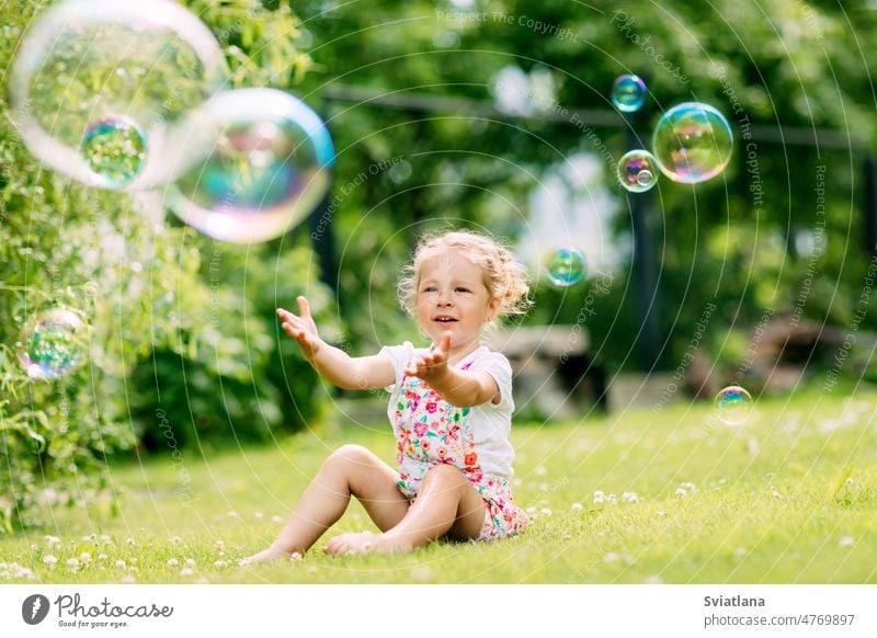 A little girl catches soap bubbles in the park, sitting on the grass. The concept of a happy childhood summer cute nature sunny kid green joy happiness small
