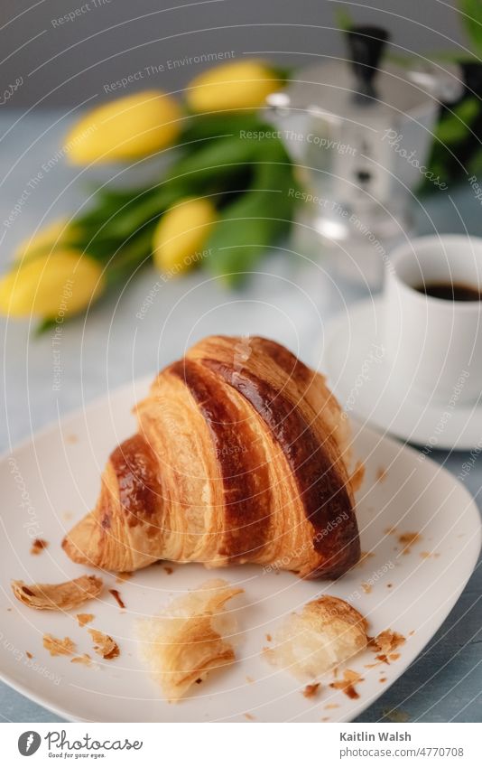 A flaky croissant sits in front of an espresso and yellow tulips. Food baked goods bakery breakfast delicious fresh eating coffee continental Flaky pastry