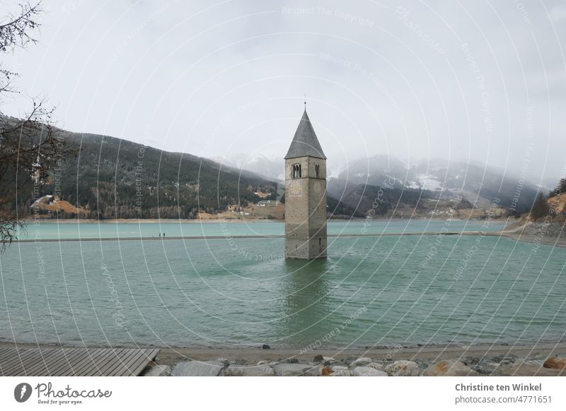 The sunken steeple of the parish church of Alt-Graun in the very empty Reschensee lake in South Tyrol and two walkers with dog on the sandbank Lake Reschen