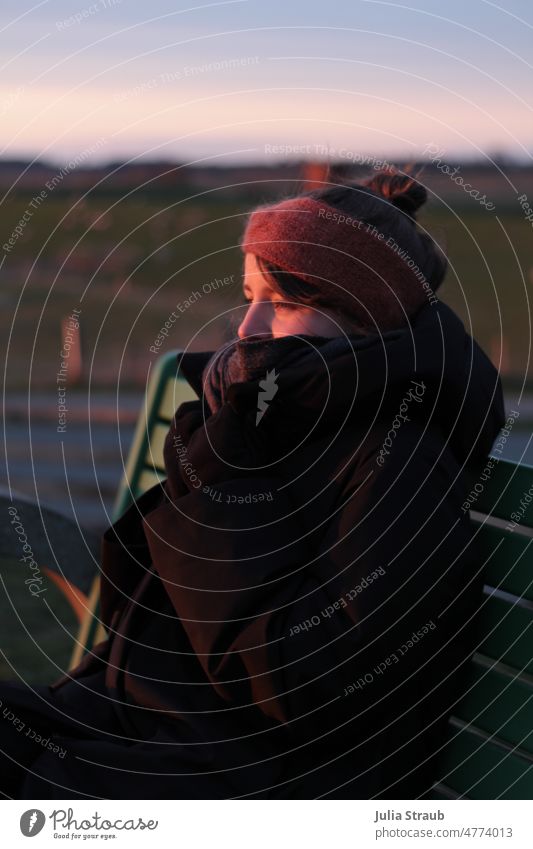 Young woman enjoying the sunset in the cold on the dike Sunlight Sunset cold season Cold North Sea Dike Green bench Headband Woman winter jacket Coat Scarf
