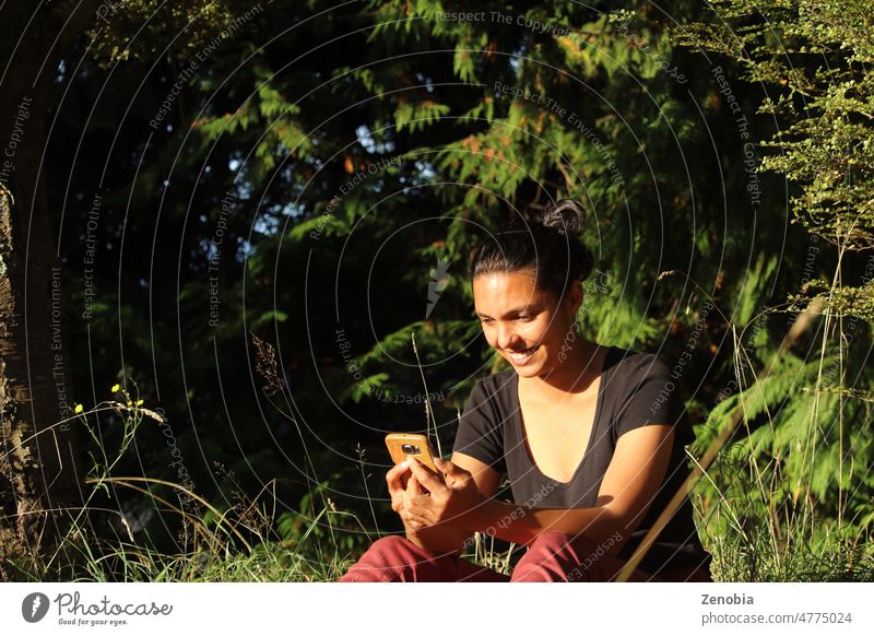 Woman sitting outside in nature, looking at phone and smiling. adult attractive beautiful black brown bun caucasian communication connection dark diverse ethnic