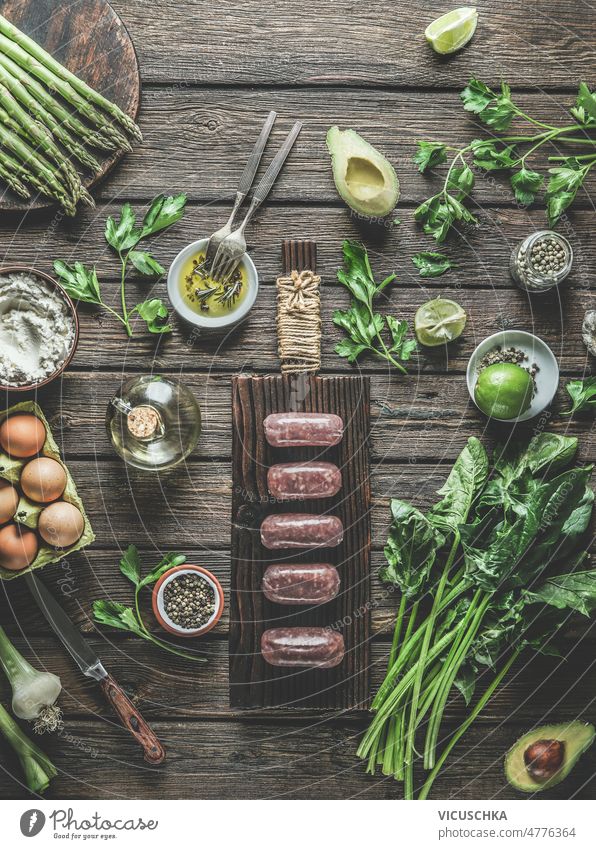 Italian sausages salsiccia on wooden cutting board at rustic brown kitchen table with various vegetables, healthy ingredients, herbs and kitchen utensils.