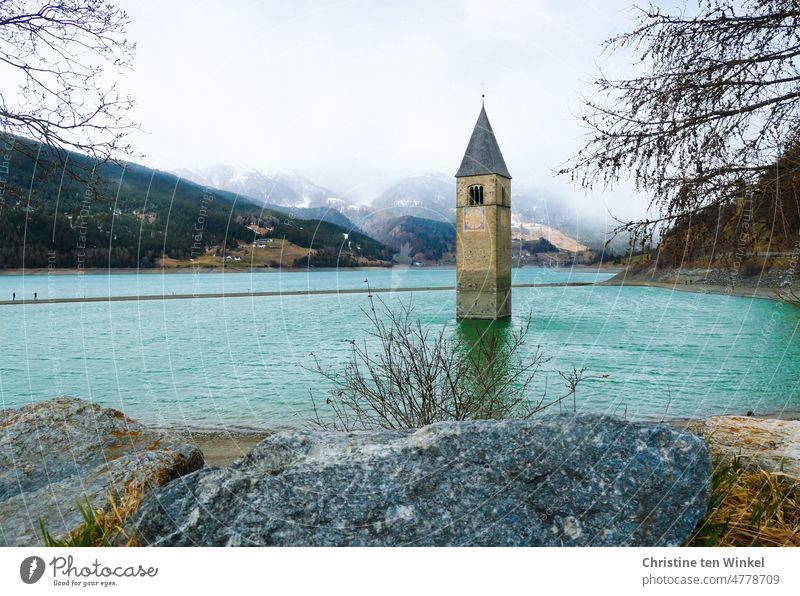 The sunken steeple of the parish church of Alt-Graun in the very empty Reschensee lake in South Tyrol. The two walkers on the sandbank are just taking a souvenir photo
