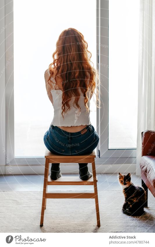 Anonymous redhead woman sitting against windows lonely solitude room living room observe style interior design curly hair domestic female apartment red hair