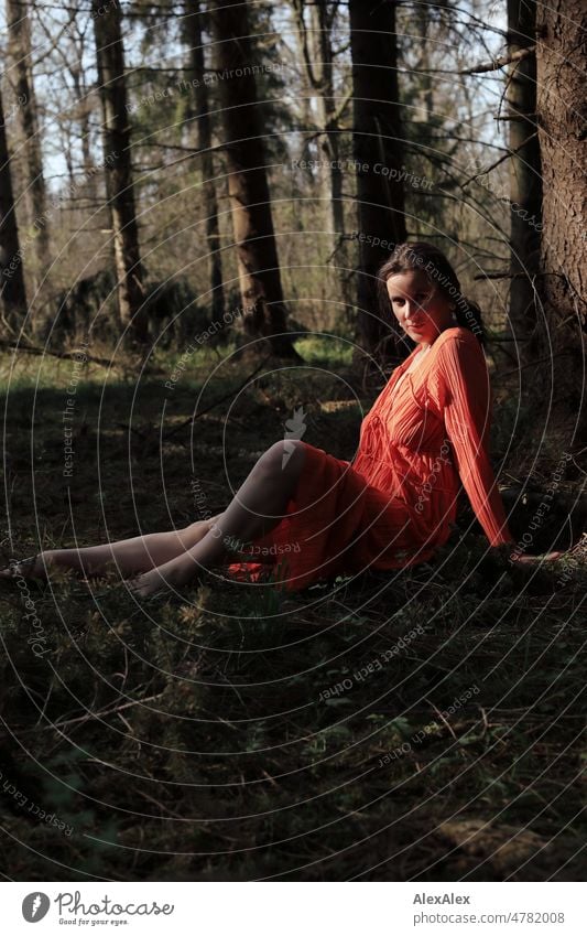 Young woman in red dress sits in spring forest and looks sideways at camera Woman youthful pretty Long-haired Brunette daintily Beauty & Beauty 18 - 30 years