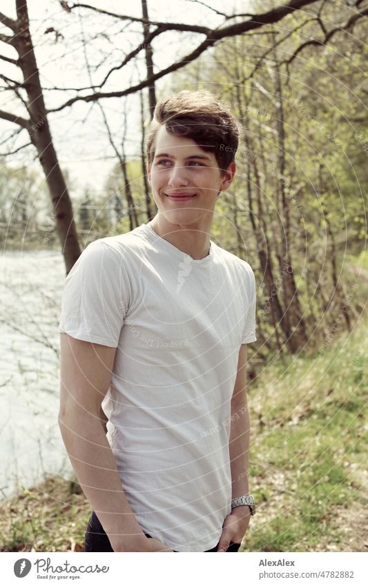 Young man standing on a lakeside and looking to the side with a smile Man younger out Nature Large pretty Athletic Dirty Blonde T-shirt white shirt kind
