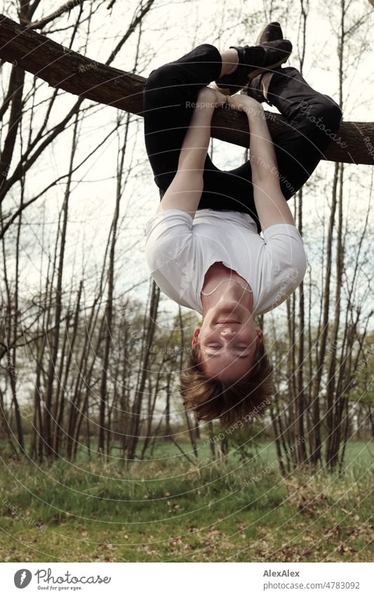 Young man hanging upside down with the back of his knees on a branch and smiling at the camera Man younger out Nature Large pretty Athletic Dirty Blonde T-shirt