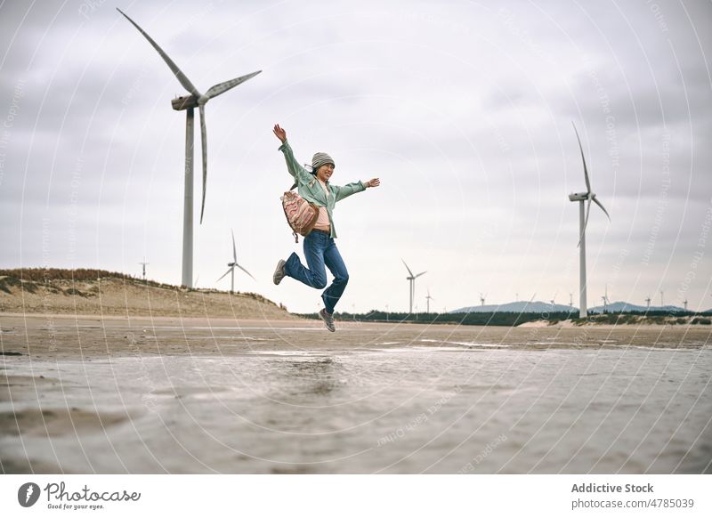 Woman jumping near windmills on seaside energy Asian girl Chinese girl cloudy beach sky freedom turbine woman weather shore water hobby outdoors full body