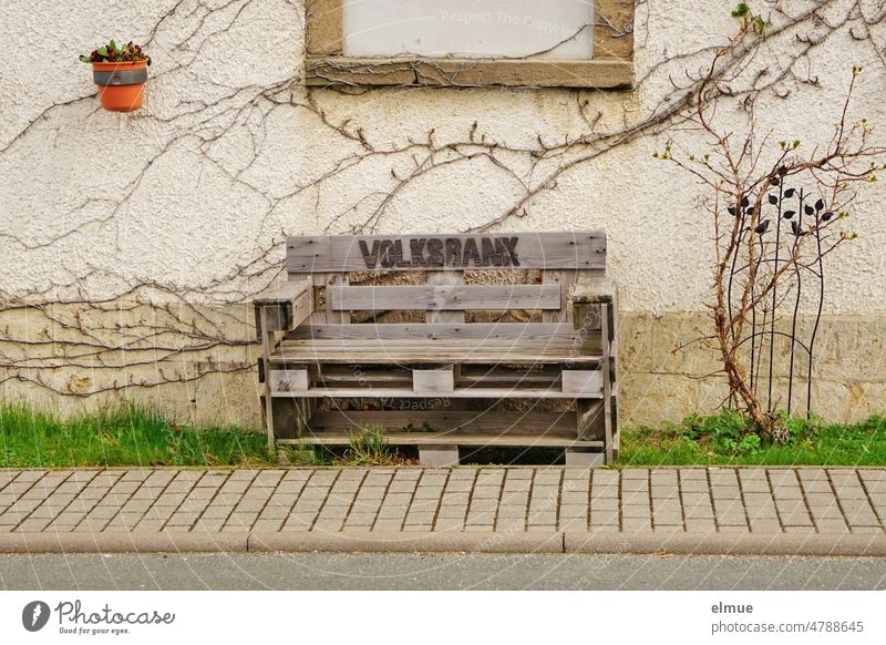 a wooden bench made of euro pallets with the inscription VOLKSBANK stands in front of a house on the lawn next to the paved sidewalk Bench Wooden bench