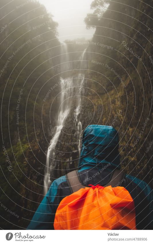 Rainy weather hiker in a waterproof jacket looks at the majestic Risco waterfall immersed in mist and rain on the island of Madeira, Portugal. Discovering magical places in Europe