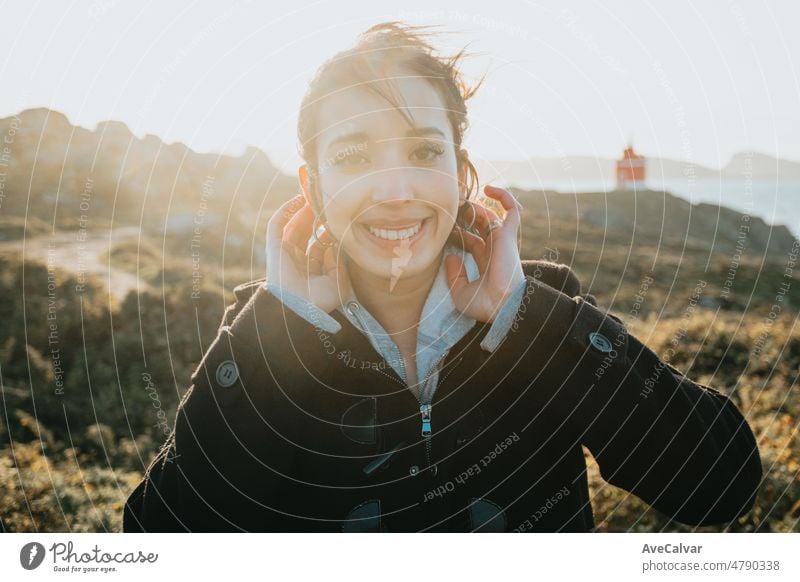 Light and vibrant portrait of a young arab woman smiling during a windy day during the sunset golden hour. Sea shore travel and holiday image, with a lighthouse behind. Van life with copy space