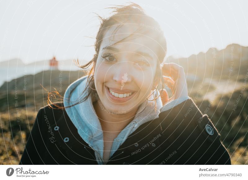 Close up Light and vibrant portrait of a young arab woman smiling during a windy day during the sunset golden hour.Sea shore travel and holiday image,with a lighthouse behind. Van life with copy space