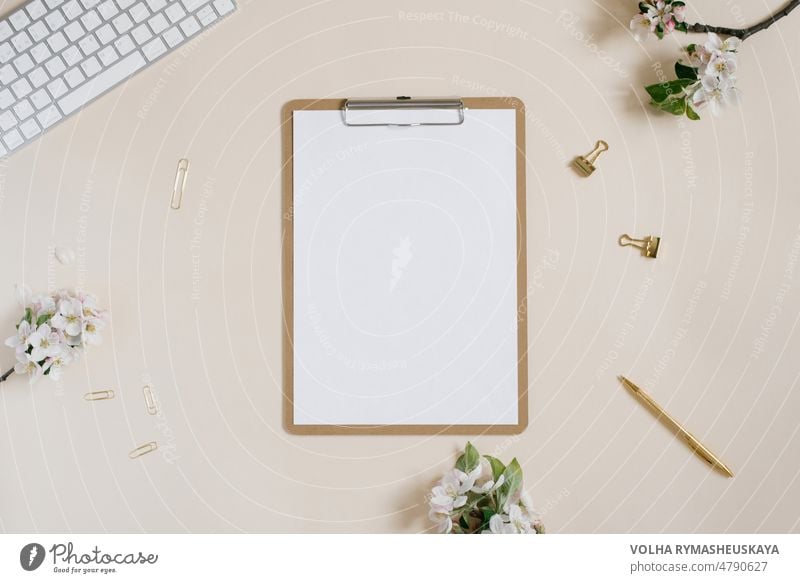 Clipboard tablet pad with blank copy space and white apple flowers, keyboard, pen and paper clips on a beige backdrop. Flat lay, top view template. Female office desk