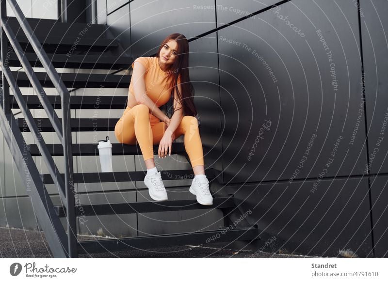 Sits on the stairs and holds bottle of water. Young woman in sportswear have fitness session outdoors female young adult body yellow clothes european caucasian