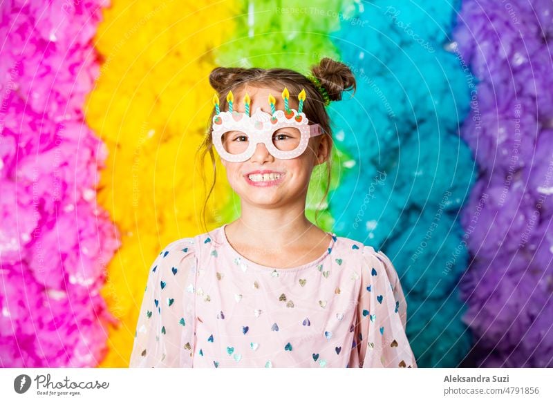 Cute girl celebrating birthday, wearing party paper glasses, laughing and having fun. Bright rainbow coloured background. Happy event. booth bright candid